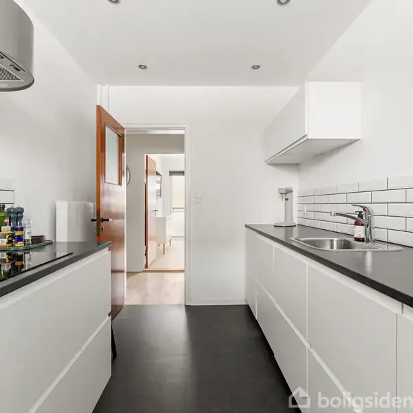 Et moderne køkken med hvide skabe og sorte bordplader. Kogeplade med emhætte på venstre side, vask på højre. Lys trædør fører til tilstødende rum.