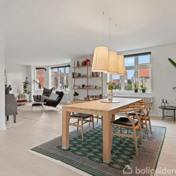 Et lyst stueområde indeholder en brændeovn til venstre, en sofa foran en fladskærm og et sofabord. Store vinduer til højre giver udsigt til omgivelserne.