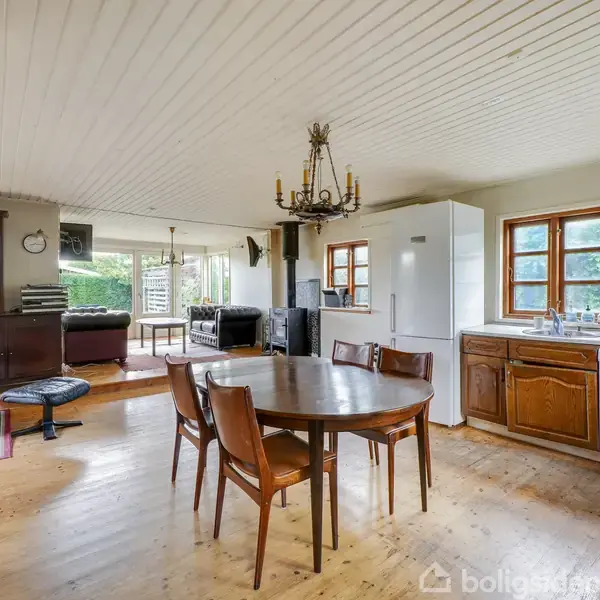 Rundt spisebord med træstole står i åben stue. Køkken med hvidevarer bagved. Trægulv og lyst træloft. Vindue ved køkkenvask, sofa og reoler i baggrunden.