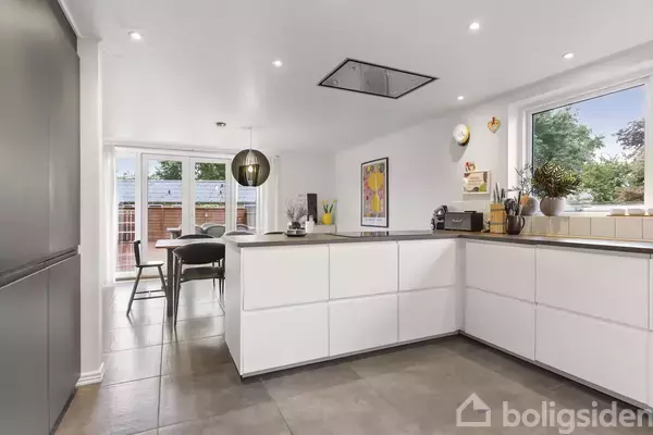 Et moderne køkken med hvide skabslåger og en central kogeø. Køkkenet åbner op til spisestue med stort vindue og glasdøre. Indretningen er minimalistisk med enkelte dekorationer.