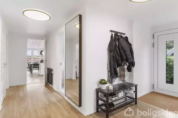 Spejl reflekterer et lyst indgangsparti med trægulv, jakker hænger på knagerække over et lille bord med hylder. Dør til højre fører ud til en have.