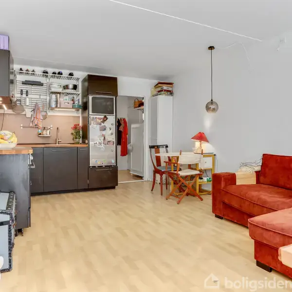 En stue kombineret med køkken, hvor en rød sofa står ved et bord i træ. Køkkenet har sorte skabe og pander, mens et køleskab står i hjørnet. En rund lampe hænger ned.
