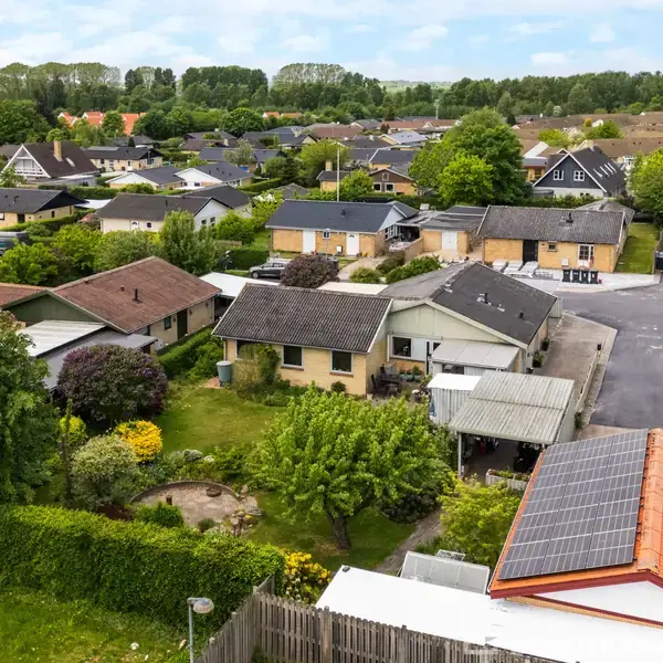 En række villaer med solpaneler på en af dem står tæt sammen, omgivet af frodige haver og grønne områder, i en dansk forstad.