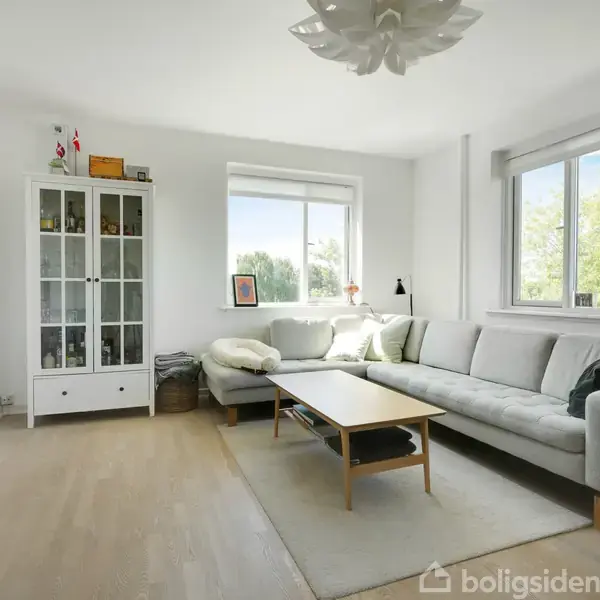 Stue med en grå hjørnesofa ved siden af vinduer, der slipper lys ind. Et sofabord står foran sofaen, og et vitrineskab står i hjørnet. Plante ved siden af sofaen.