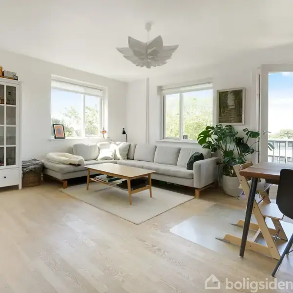 Stue med en lys grå sofa placeret ved vinduerne, der giver udsigt til en altan. En hvid vitrineskab står til venstre, mens et spisebord med stole står til højre.