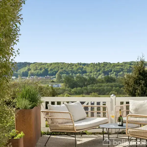 En terrasse med hvide kurvemøbler og puder står stille. Konteksten omfatter en frodig, grøn skov i baggrunden og en klar blå himmel, der skaber en rolig scene.