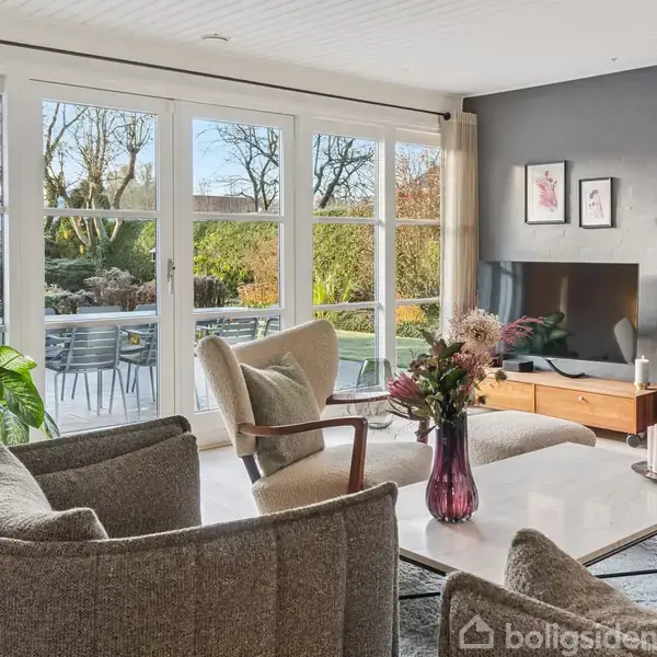 En stue med kurvet sofa og lænestol omkring et sofabord, hvor en vase med blomster står. I baggrunden ses store vinduer, græsplæne og terrasse med havemøbler. En brændeovn står ved siden af et TV på en træbænk.