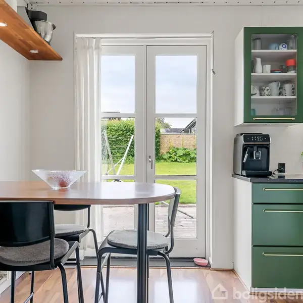 Et køkkenbord med to stole står foran en glasdør, der åbner ud til en have. Til højre ses grønne køkkenskabe og en kaffemaskine.