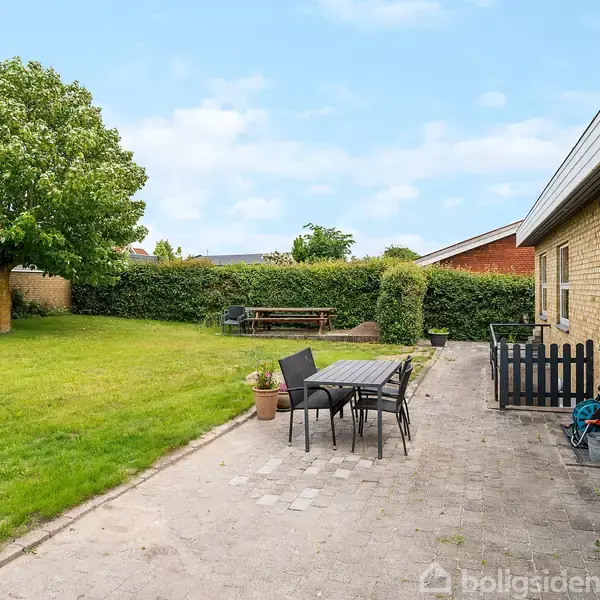 Terrasse med havebord og stole står på stenterrasse ved gult murstenshus. Græsplæne med træ og hæk i baggrunden skaber en indbydende, grøn haveatmosfære.