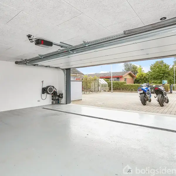 En tom garage med åben port, udsigt til en indkørsel og to motorcykler parkeret udenfor. Hvide vægge, glat gulv, og udstyr hængende på væggen.