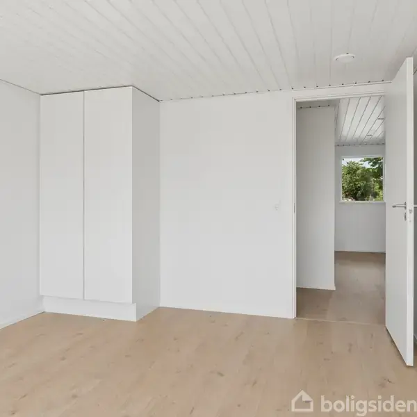 Et tomt rum med hvidmalede vægge og et lyst trægulv. Et indbygget skab står til venstre. En åben dør fører til en anden del af huset, hvor man kan se et vindue.