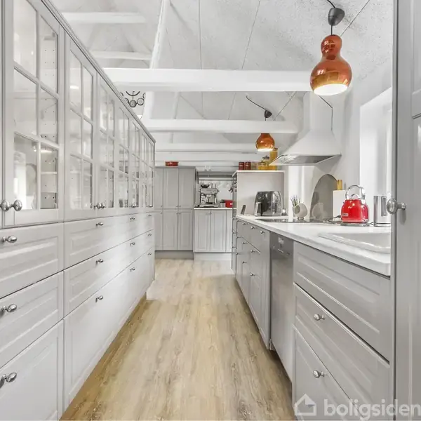 Et lyst, moderne køkken med grå skabe og trælaminatgulv. Orange lamper hænger over køkkenbordet, hvor der står en rød tekedel og kaffemaskine. Loftet har synlige bjælker.