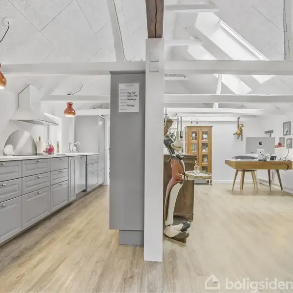 Køkken med grå skabe og ovne, lyst trægulv under loftsbjælker, åbner til stue med bogreol, skrivebord, stole, og lyse vægge. Tekst på skab: "Thank you for keeping this place nice and tidy".