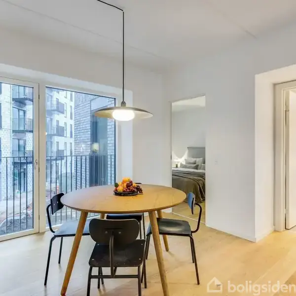 Et rundt spisebord med fire stole står i en lys stue. En lampe hænger over bordet. Store vinduer viser en balkon og udenforliggende bygninger. Til højre ses et rum med vaskemaskine og tørretumbler.
