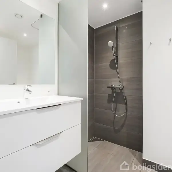 Badeværelse med hvid vask og skab, brusekabine med bruser på væggen, omgivet af mørke fliser, i en moderne og minimalistisk stil.