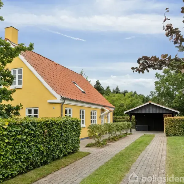 Gult hus med rødt tag står i en grøn have. En flisebelagt sti fører til en sort carport, omgivet af hække og træer.