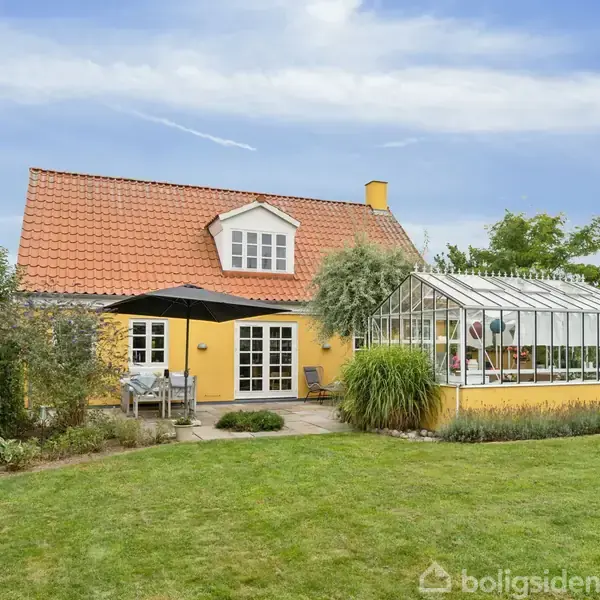 Gult hus med rødt tegltag står i en frodig have. Ved siden af ses et stort drivhus, og foran huset er en terrasse med parasol og havemøbler.