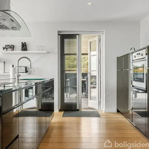 Moderne køkken med sorte højglansskabe og indbygget ovn; glasdør fører ud til terrasse med grøn udsigt; lyse trægulve og stilrent design skaber en lys og åben atmosfære.