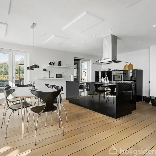 Et moderne køkken med spisebord og stole. Ved siden af står en brændeovn i hjørnet. Store vinduer giver naturligt lys, og rummet har minimalistisk indretning.