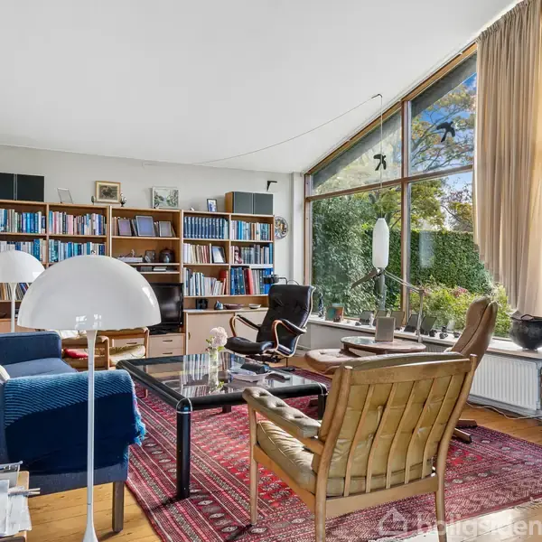 En stue har møbler som sofaer og stole omkring et sofabord. Store vinduer giver udsigt til grønne omgivelser. En bogreol fyldt med bøger og kunst dekorerer rummets vægge.