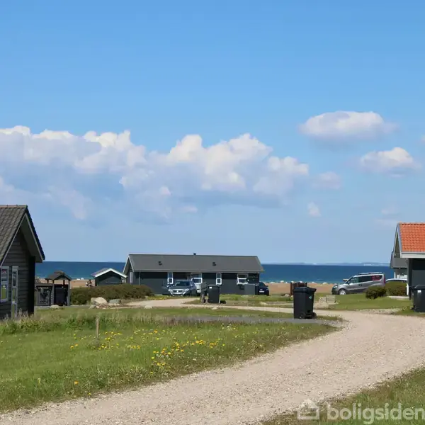 Flere små huse står spredt i et sommerhusområde. En snoet grusvej løber gennem de grønne græsarealer. I baggrunden ses havet under en blå, skyet himmel.