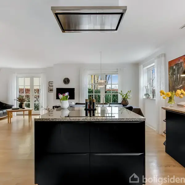 Sort køkkenø med marmorbordplade står i et moderne, lyst køkken-alrum. Køkkenet har hængende lamper, planter, og træudskæring på væggen. Stue med sofa og tv ses i baggrunden.