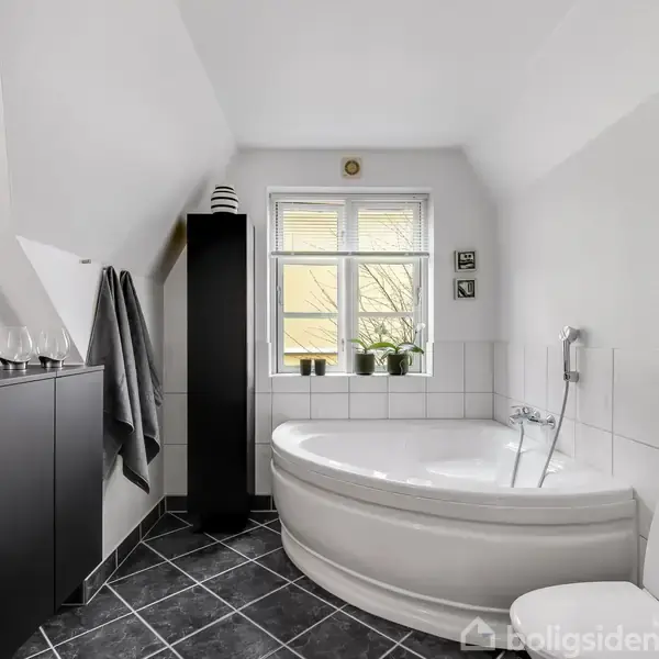 Et moderne badeværelse med et rundt badekar i hjørnet, flankeret af to planter på vindueskarmen. Der er sorte skabe og grå håndklæder, alt i et lyst, flisebelagt rum.
