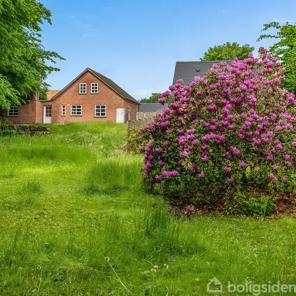 Rødt murstenshus står stille i frodig have, omgivet af grønne træer og en stor busk med lyserøde blomster.