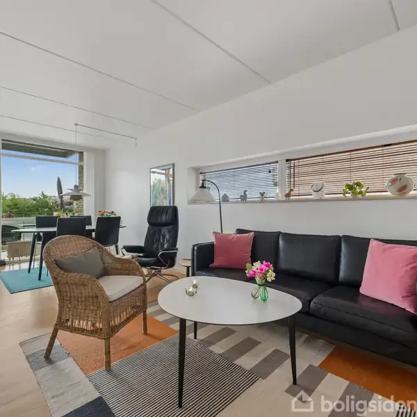 Stue med sort sofa, to farvede puder og et rundt bord foran. Til venstre en fletstol og spisebord med stole nær glasdør, som åbner til en terrasse med udsigt over have.