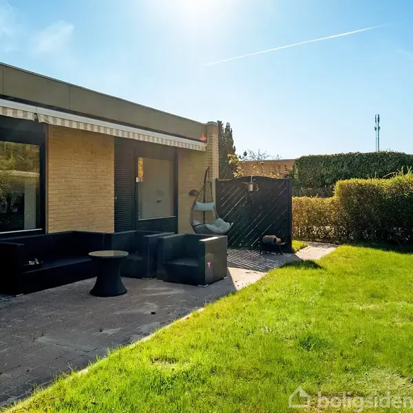 Terrasse med sorte møbler under en markise, beliggende ved et murstenshus. En velplejet græsplæne omgiver området, indrammet af hække under en klar, blå himmel.