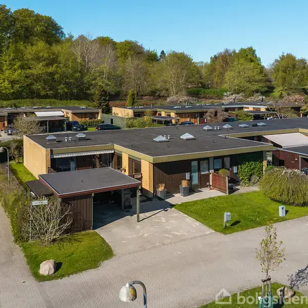 Fladt taghus i moderne kvarter med grønne områder i baggrunden. Omgivet af lignende bygninger og vegetation. Fotograferet fra luften under en klar himmel.