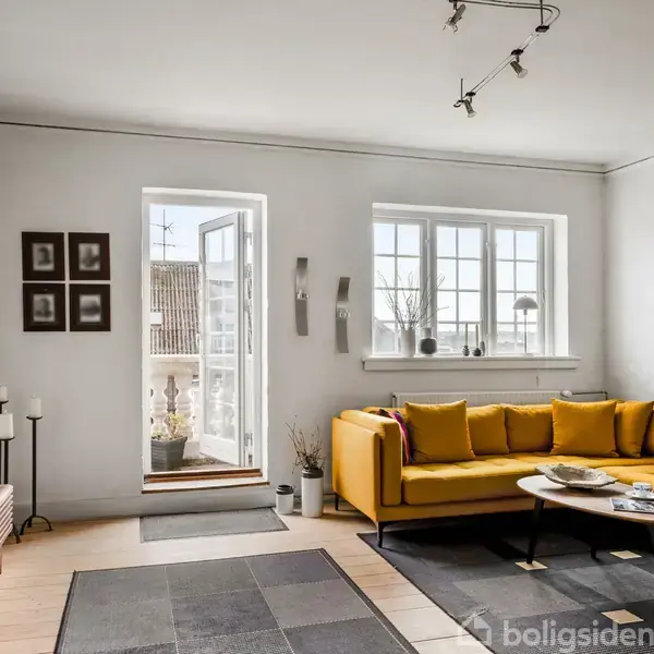 En gul hjørnesofa står i et lyst stueområde. Sofaen er placeret foran store vinduer og en åben dør til en balkon. Væggene er dekoreret med kunst, og der er et stort fladskærms-tv på møbel enhed.