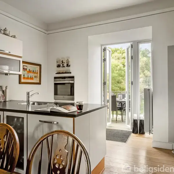 Køkken med ø, hvorpå der står et glas rødvin og åbne bøger; til højre et køleskab. Gennem åbne døre ses en have med træer og havemøbler.