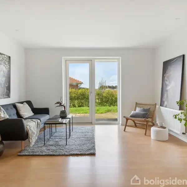 Sofa med tæppe står i lys stue med hvide vægge. Store vinduer viser grøn have udenfor. To billeder hænger på væggene; en stol og plante dekorerer rummet.