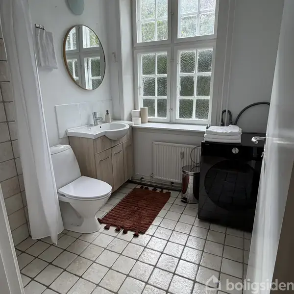 Toilet placeret ved siden af hvid vask, omgivet af skab. I nærheden står en vaskemaskine. Rød badeværelsesmatte på flisegulv, vindue i baggrunden med spejl på væggen.