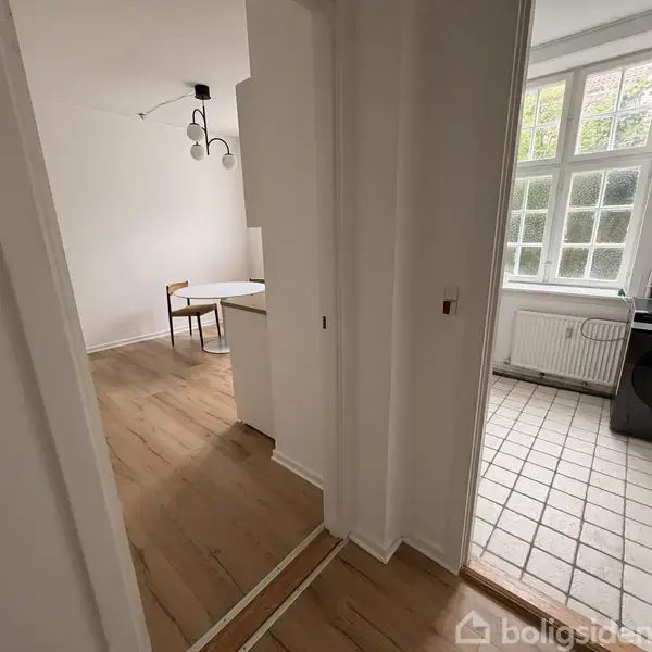 Døråbning forbinder køkken med spisestue. I spisestuen står et rundt bord med to stole. Køkkenet har flisegulv, stort vindue og vaskemaskine ved siden af en radiator.