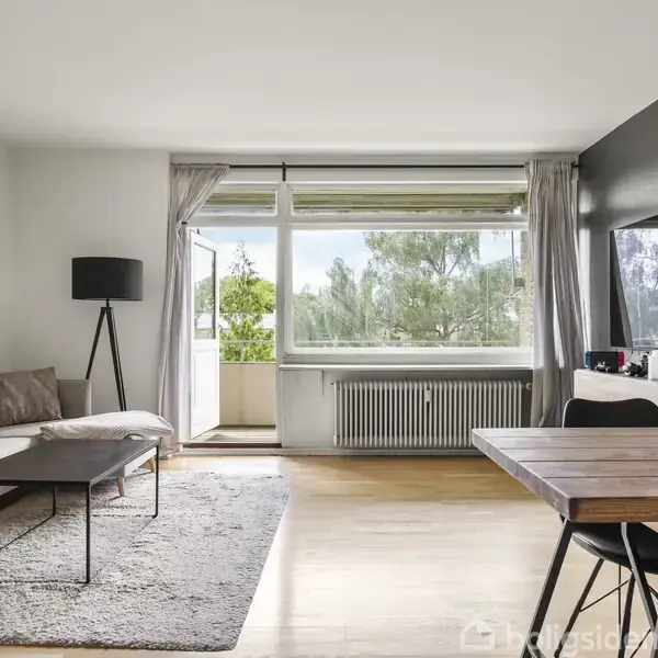 Sofa står i en lys stue med et tæppe og et sofabord foran. Store vinduer og en døren åbner ud til en balkon med træer i baggrunden.