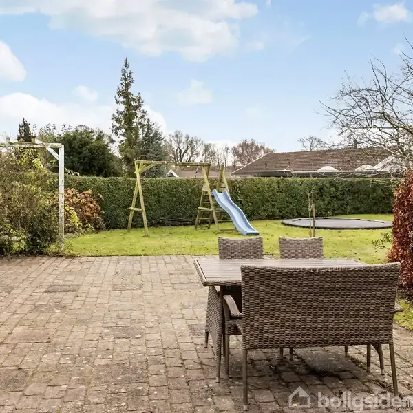 Havegruppe med flisebelagt terrasse, hvor et bord med to stole står. I baggrunden er der en legeplads med en gynge og en rutsjebane på en græsplæne.