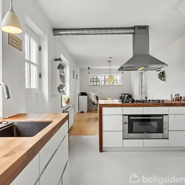 Et moderne køkken med hvide skabe og en træbordplade. En vask er placeret til venstre, mens en kogeø med indbygget ovn og emhætte er centralt, omgivet af lyse vægge.