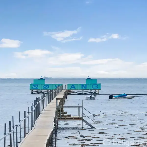 En lang træbro fører til en badeanstalt med skiltet "SØBAD". Broen strækker sig over havet, omgivet af lavt vand. I baggrunden ses både og en klar himmel.