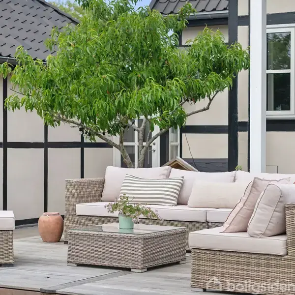 Sofaarrangement med lyse puder står på en terrasse af træ. Et lille bord med en plante foran, mens et grønt træ og sort-hvid bindingsværkhus udgør baggrunden.