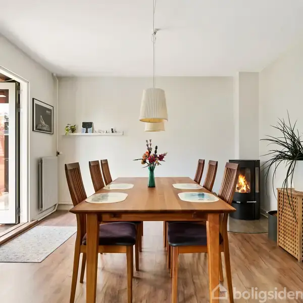Et træspisebord med seks stole er opstillet i en lys spisestue. Bordet er pyntet med en vase med blomster, og i baggrunden ses en brændeovn og en stor plante.