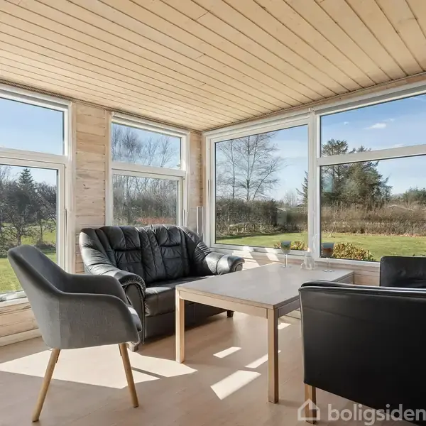 Stue med to sorte lænestole og en grå stol omkring et træbord. Store vinduer giver udsigt til en grøn have med buske og træer under en blå himmel.