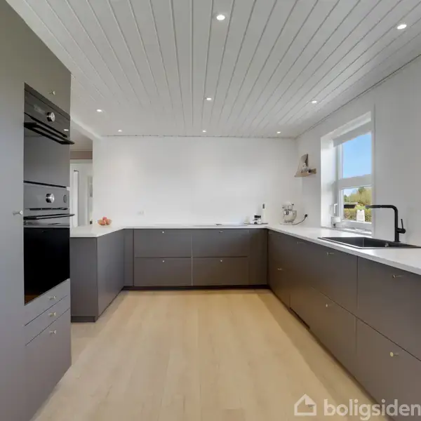 Køkken med grå skabe og indbygget ovn; rummeligt og moderne design; lyst trælaminatgulv og hvidt loft med spots; stort vindue til højre giver naturligt lys.