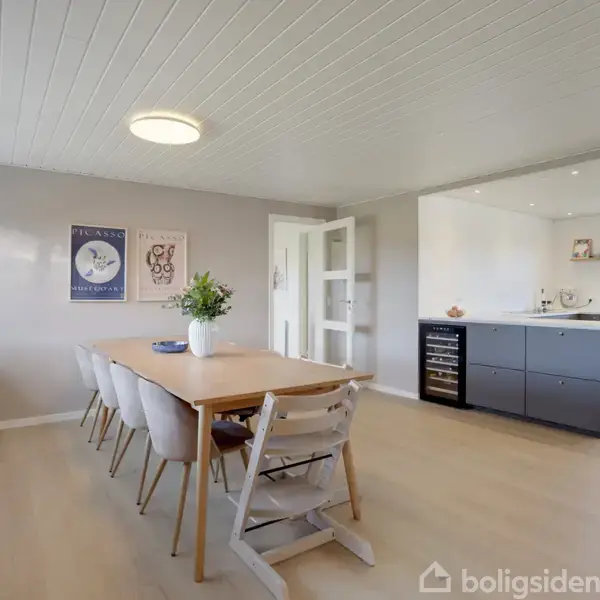 Spisebord med stole står i en lys stue, hvor en enkel plante pynter bordet. Køkkenet ses i baggrunden. Væggen har to kunstplakater, én med navnet "Picasso."