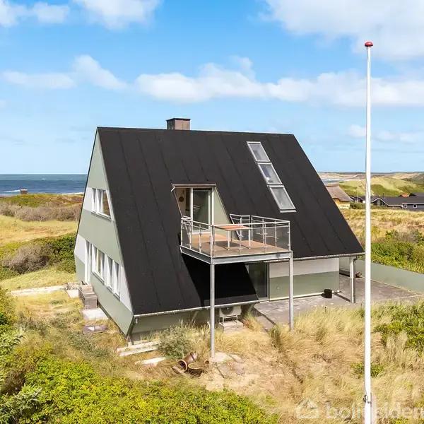 Et moderne hus med trekantet tag har en balkon og ligger i klitlandskab tæt ved stranden. Blå himmel og hav ses i baggrunden, omgivet af græs og buske.