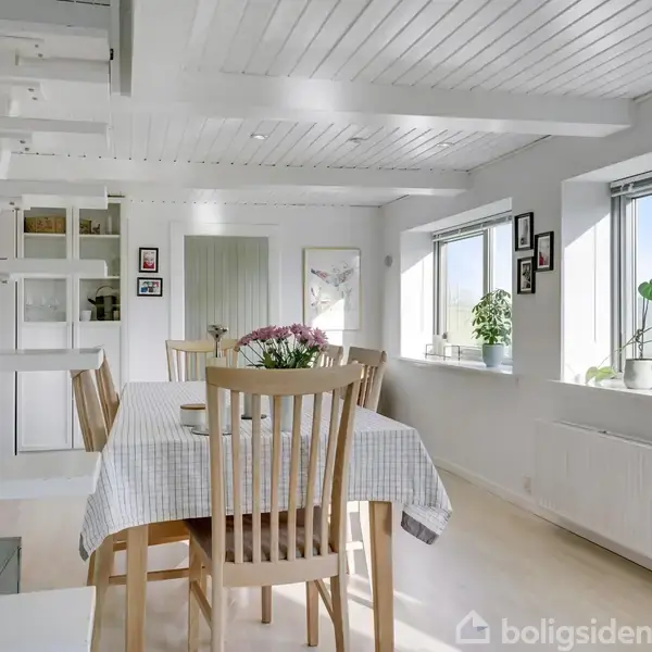 Spisebord omgivet af træstole står i et lyst køkken-alrum. En trappe fører til en hems, mens vinduerne fylder rummet med lys. Blomster og dekorationer pynter rummet.