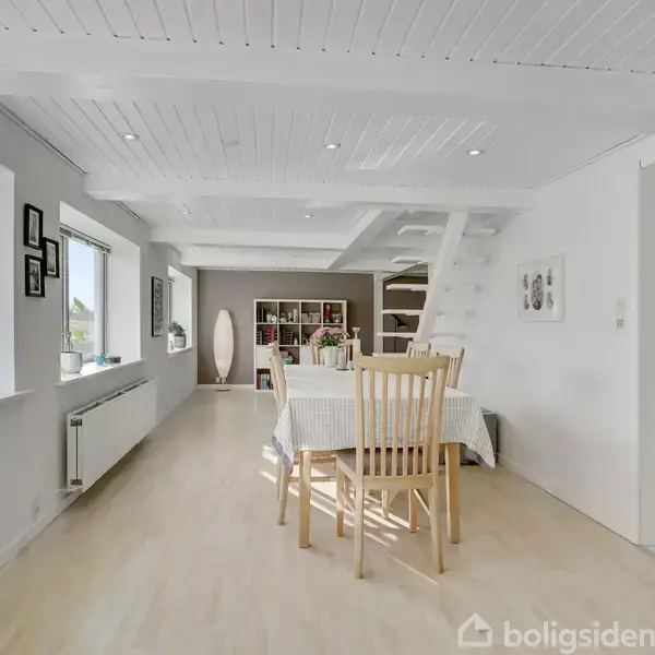 Spisebord med træstole under en hvid, trækonstruktion i et lyst rum med trægulv. Til venstre ses vinduer med planter og billeder, og til højre et åbent køkkenområde.