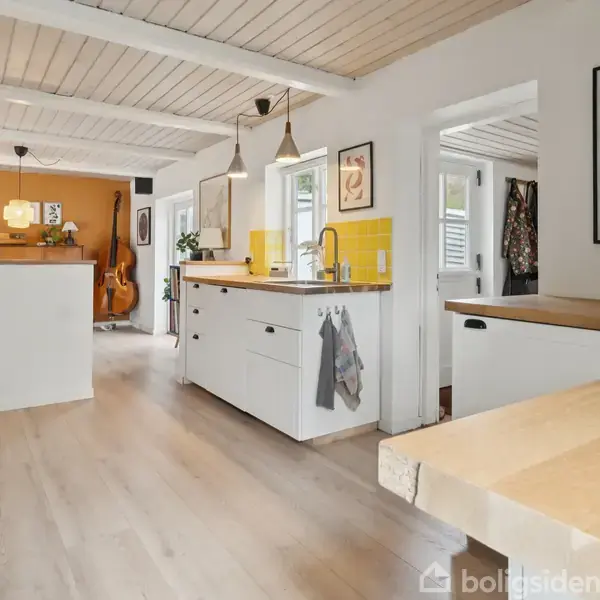 Et lyst køkken med hvide skabe og træbordplader. To lamper hænger over en køkkenø. En farverig væg med malerier. I baggrunden står en kontrabas. Ingen tekst fundet.