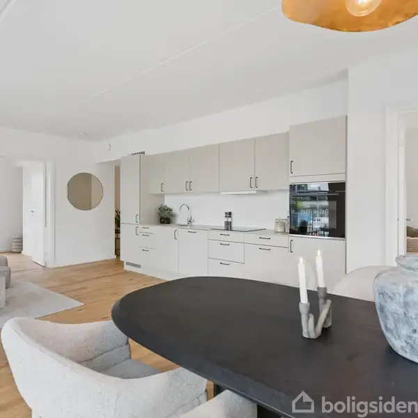 Et moderne køkken og stue i åben planløsning med hvide skabe og træbord. En grå sofa står mod væggen, og der er en dør til et soveværelse i baggrunden.